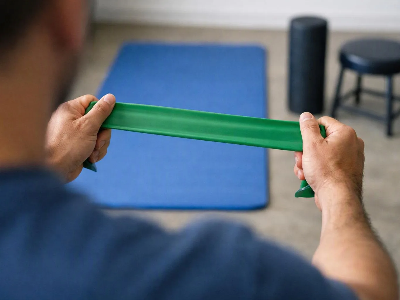 A person stretches a green resistance band over a blue exercise mat in a gym room