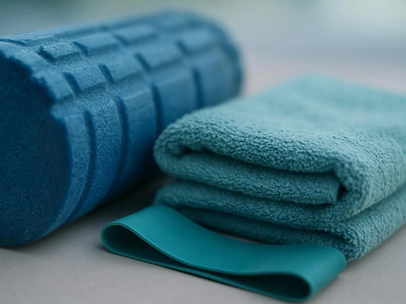 A rolled blue foam mat, folded teal towel, and teal exercise band rest on a light surface
